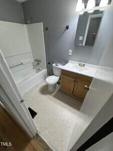 Small bathroom with white bathtub and shower, toilet, wooden vanity with sink, wall mirror with lights, and light gray walls. The floor has light-colored tiles, and part of a wooden floor is visible just outside the door.