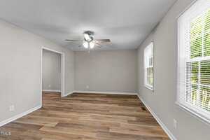 A nearly empty room with light gray walls, wood-look flooring, a ceiling fan with lights, and several windows with white blinds letting in natural light. An open doorway leads to another room with similar flooring.