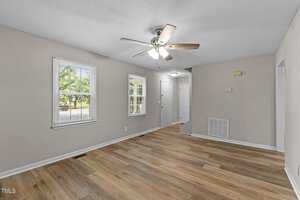 A bright, empty room with light wood flooring, beige walls, two windows, a white front door, a white interior door, a ceiling fan with lights, and a vent cover on the wall.
