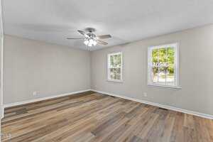 Empty room with light-colored walls, wood-style flooring, a ceiling fan with lights, and two windows letting in natural light. The windows show some greenery outside.