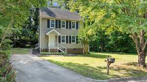A two-story beige house with black shutters and a covered front porch, surrounded by grass and mature trees. A concrete driveway leads to a mailbox at the edge of the front yard.