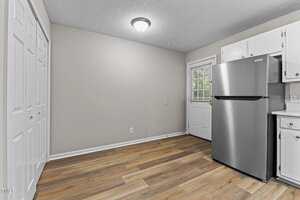 A kitchen with light wood flooring, white walls, white cabinets, a stainless steel refrigerator, and a door with a window leading outside. A ceiling light fixture is centered on the textured ceiling.