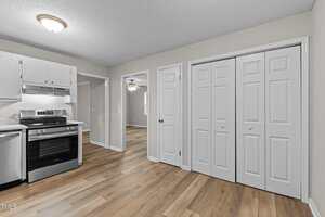 A kitchen with light-colored walls, white cabinets, a stainless steel stove and dishwasher, wood-style flooring, and two sets of white double doors. An open doorway leads to another room with a ceiling fan.