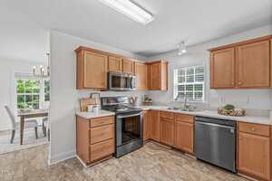 A modern kitchen with wooden cabinets, stainless steel appliances including a stove, microwave, and dishwasher, and a window above the sink. Adjacent dining area with a window and chandelier is partially visible.