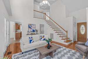 A living room with a white sofa, a wooden coffee table, and a patterned rug. A staircase with wooden steps and a white railing leads to an upper level. A wooden front door is at the right side of the image.