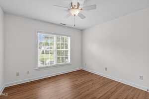 A bright, empty room with white walls, a ceiling fan with a light, a large window with blinds, and wood-look flooring. Electrical outlets line the walls and sunlight enters through the window.
