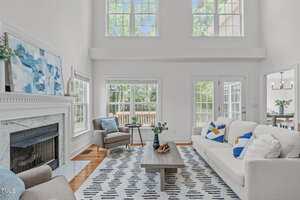 A bright living room with tall windows, white walls, a white sofa with blue and yellow pillows, two gray chairs, a fireplace, a patterned rug, and a coffee table. Dining area visible through an open doorway.