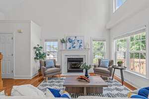 A bright living room with two gray armchairs, a wooden coffee table, and a white fireplace. Large windows let in sunlight. The room features a patterned rug, potted plants, and modern decor.