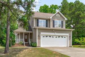 Charming two-story beige home at 2575 Primrose Lane in Creedmoor with a covered front porch, attached two-car garage, white trim, multiple windows, asphalt driveway, brick steps, and lush trees and greenery all around.