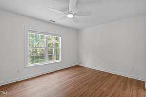 An empty room with white walls, a large window letting in natural light, a ceiling fan with a light, and wood-style flooring. Trees are visible outside the window.