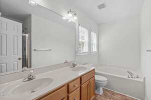 A bathroom with a double sink vanity, wood cabinets, a large mirror, a bathtub, a toilet, white walls, and tile flooring. Two windows with blinds let in natural light.