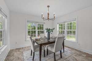 A dining room with a wooden table set for four, light gray upholstered chairs, a central modern chandelier, and two large windows letting in natural light. The floor is tiled, and the walls are painted white.