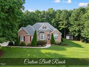A large brick ranch-style house at 517 Belmont Circle, Wake Forest, with a manicured lawn, bushes, and trees surrounding the property. The home features multiple gables and white-trimmed windows. "Custom Built Ranch" text is overlaid at the bottom.