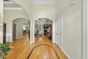 A hallway with wood floors and cream-colored walls leads to a living area with a fireplace. Multiple arched doorways and a white door are visible, along with a plant on the left. Ceiling fan and furniture in the background.