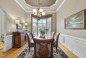 A formal dining room with a wooden table and six chairs, a sideboard with two lamps, a chandelier, a plant centerpiece, an area rug, wainscoting, a large window with shutters, and a framed ocean painting.
