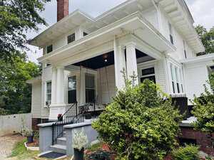 A two-story white house with a covered front porch supported by large columns, black railings, and several potted plants near the steps. A white fence and greenery are visible in the background.