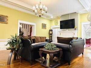 A living room with yellow walls, brown leather sofas, a glass coffee table with a plant, hardwood floors, a chandelier, and a fireplace with a TV mounted above it. Large windows provide natural light.
