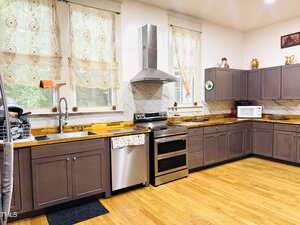A spacious kitchen with brown cabinets, wood countertops, stainless steel appliances, lace curtains, and hardwood floors. Decorative items and small appliances are placed on the counters and upper cabinets.