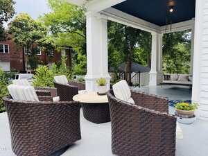 A covered porch with four brown wicker chairs around a round table, potted plants, and a swing hanging in the background, surrounded by white columns and greenery.