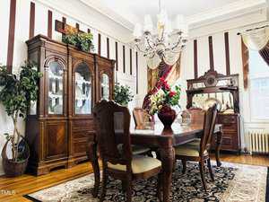 A formal dining room with a wooden table and six chairs, a china cabinet, a sideboard, a chandelier, large windows with drapes, indoor plants, and a decorative rug on a hardwood floor.