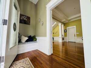 A view from an open doorway into a hallway with hardwood floors, white trim, pale green walls, and decorative wall art. A built-in bench with pillows is visible on the left side of the entry area.