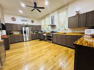 Spacious kitchen with dark brown cabinets, wood countertops, stainless steel appliances, light wood floors, ceiling fan, and decorative items on the counters and above the cabinets. Two large windows have lace curtains.