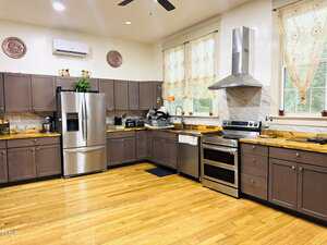 A spacious kitchen with wooden floors, brown cabinets, stainless steel appliances, a double oven, a fridge, a range hood, and countertops with kitchenware. Two large windows have lace curtains.