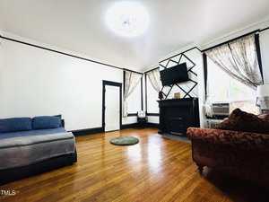 A living room with hardwood floors, a blue-gray couch, a red patterned armchair, a black fireplace with a TV above it, sheer curtains on tall windows, and a small round green rug in the center.