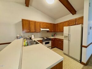 A kitchen with wooden cabinets, white countertops, a white refrigerator, a stove with an overhead vent, a double sink, and cleaning supplies on the counter. Exposed wood beams are visible on the ceiling.