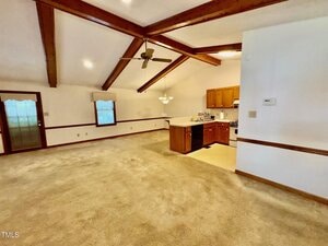Spacious open-plan living area with vaulted ceiling, exposed wooden beams, carpeted floor, and an adjacent kitchen featuring wood cabinets, a gas stove, and a ceiling fan. Large window and door let in natural light.