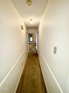 A long, narrow hallway with beige carpet and white walls, lit by two ceiling lights, with doorways on the left and right and a room visible at the end.