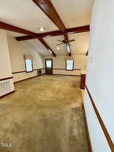 A carpeted living room with exposed wooden ceiling beams, white walls with wood trim, a ceiling fan, several windows, and a door leading outside. Built-in shelves and vents are visible along the walls.