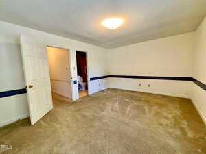 Empty carpeted room with beige walls, a ceiling light, open door leading to a bathroom with a visible toilet, and a dark blue decorative border running around the walls at mid-height.