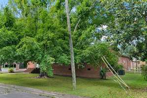 A wooden utility pole leaning to the right with wires hanging low, positioned in front of a brick house surrounded by green trees and grass on a suburban street.