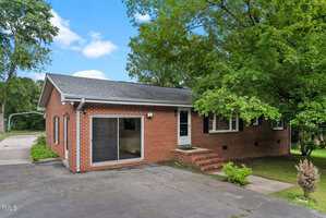 A single-story red brick house at 221 W Dale Drive, Oxford, features a gray shingle roof, large front window, small porch with steps, and a paved driveway. A tree partially shades the green grass that surrounds the house.