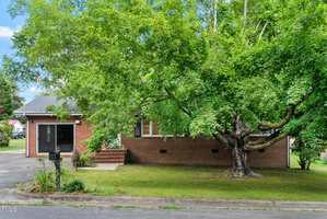 A single-story brick house with a large leafy tree in the front yard, a mailbox by the driveway, and a small set of steps leading to the main entrance. The house has a grassy lawn and is partially shaded by the tree.