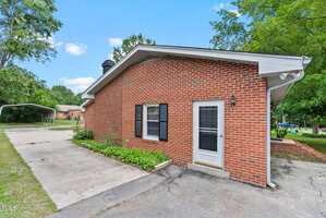 A red brick house with a white-framed door and black shutters sits next to a concrete driveway. Trees and greenery surround the property, and a carport is visible in the background. The sky is clear and blue.