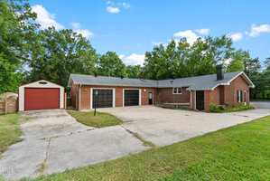 Single-story brick house with three black garage doors, a red shed with a roll-up door, a large concrete driveway, and surrounding trees under a blue sky.