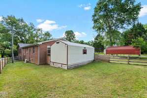 A single-story brick house with an attached white shed, surrounded by a grassy yard and wooden fencing. There are trees and a red-roofed structure in the background under a blue sky.