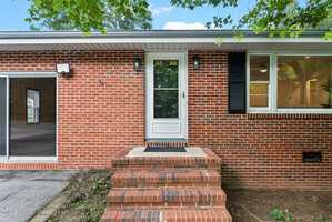 A red brick house exterior with a white storm door, black shutters, and three brick steps leading to the entrance; a window and a small light fixture are beside the door, with a tree partially shading the scene.