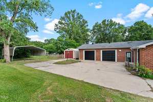Concrete driveway leading to a brick house with two black garage doors, a red shed, a metal carport, and green grass with trees under a partly cloudy sky.