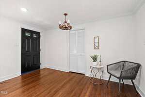 A small entryway with hardwood floors, a black front door, a white folding closet door, a modern black chair, a small white side table with a plant, and a wall mirror. A brown chandelier hangs from the ceiling.