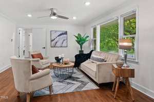 A modern living room with two beige armchairs, a matching sofa, a round wooden coffee table, a geometric rug, a side table with a lamp, a large plant, wall art, hardwood floors, and large windows with blinds.