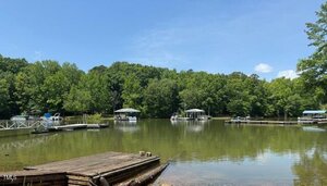 A calm lake with several boat docks and boats along the shore, surrounded by dense green trees under a clear blue sky. A wooden dock extends into the water in the foreground.