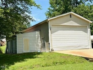 A light beige detached garage with a large white roll-up door and an attached side shed. The building sits on a grassy lawn with a concrete driveway, surrounded by trees under a blue sky.