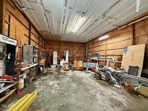 A large, cluttered garage or workshop with various tools, equipment, and materials scattered around. The concrete floor has stains and debris, and the wooden walls show exposed framing. The ceiling is unfinished with visible seams.