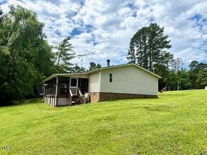 Single-story house with light-colored siding and a brick foundation on a grassy slope. The house has a screened porch, outdoor chairs, and is surrounded by trees under a partly cloudy sky. A swing set is visible in the background.