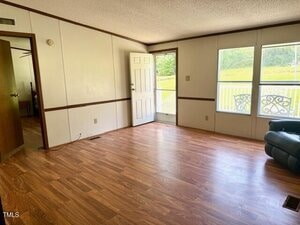 A bright, empty living room with wood-style flooring, white walls with brown trim, large windows, an open door, and a leather armchair partially visible in one corner. A porch railing and grass are visible outside.