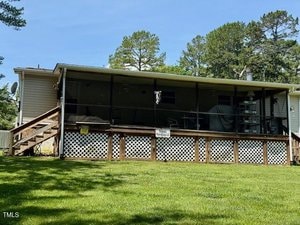 A single-story house with a screened-in back porch, wooden steps, and a lattice skirt. The yard is grassy, and tall trees are visible in the background under a blue sky.