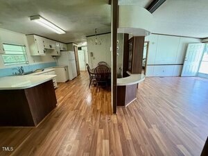 An open interior features a kitchen with white cabinets and appliances, a dining area with a wooden table and chairs, and a living space with wood flooring and natural light from windows and a glass door.